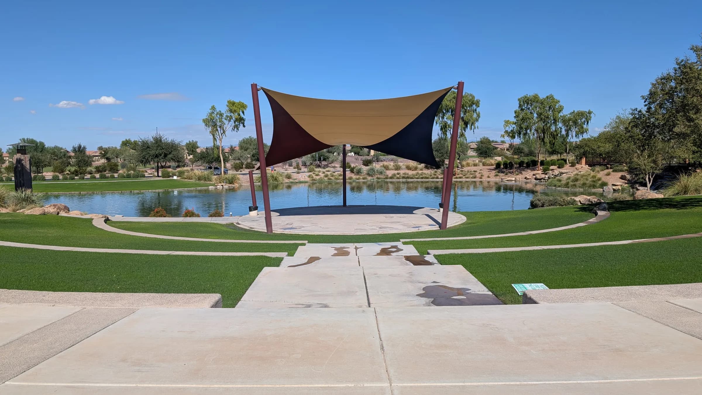 A stage under a shade sail by a pond at Anthem at Merrill Ranch, with a terraced lawn in front and desert trees behind