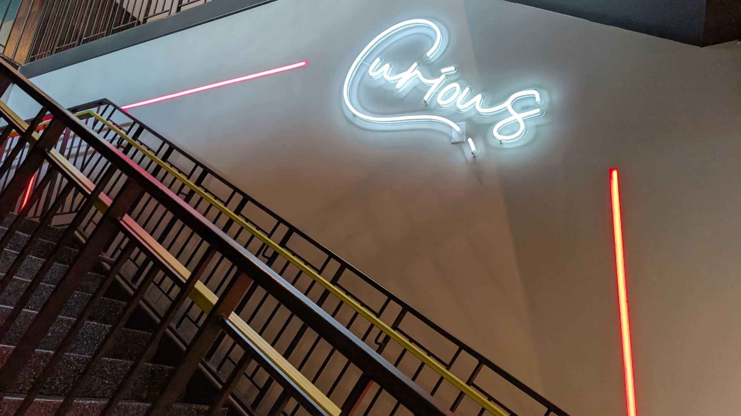A white neon sign that reads "Curious" above a stairwell at Hotel Celare, with thin red light lines on the wall