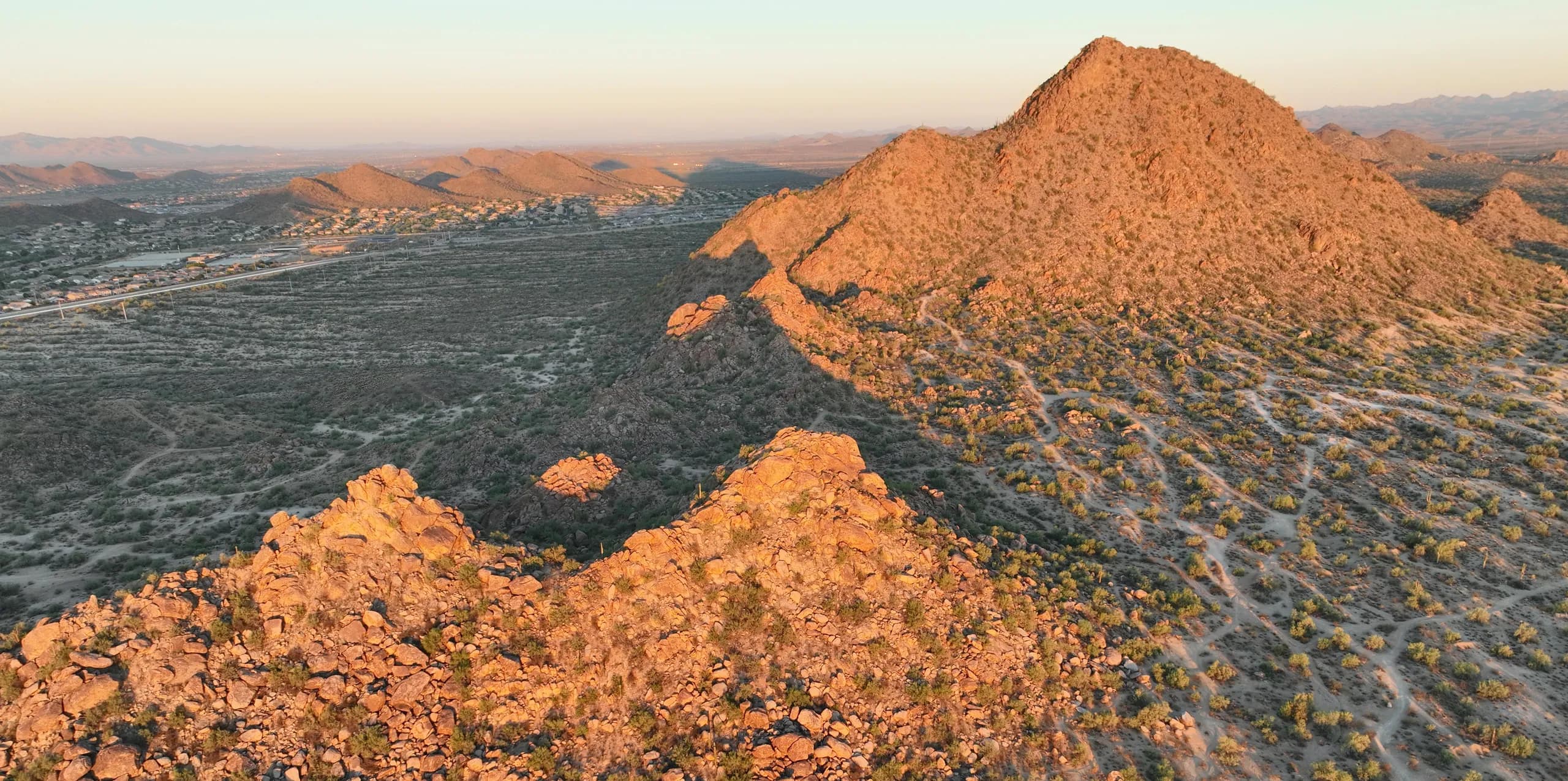The North Phoenix desert from the air at golden hour, with boulder-covered hills, dirt tracks across the flats below and neighborhoods in the distance