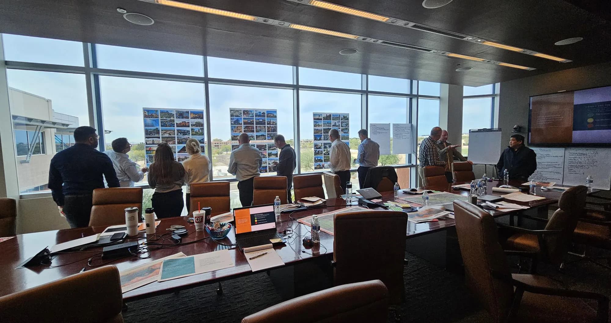 People standing at boards of images along a window wall during the NorthPark workshop, with laptops and notes on a long conference table