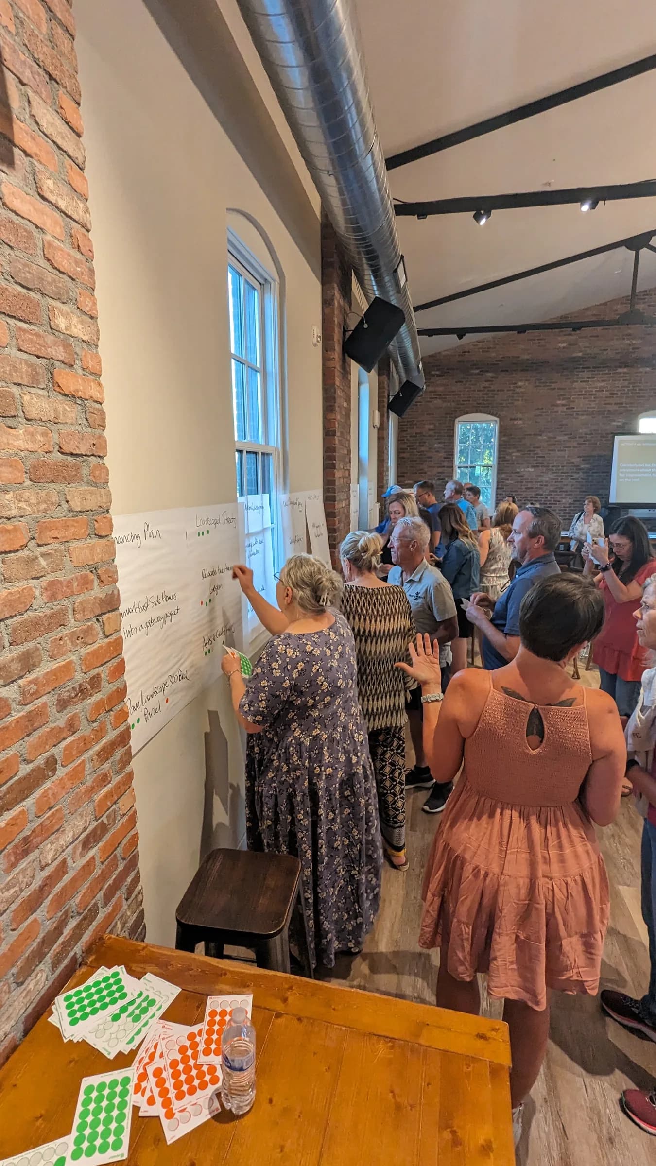 Windsor residents lining up to add their ideas to a paper wall at a downtown planning workshop, with sheets of green and orange dot stickers on a table in front