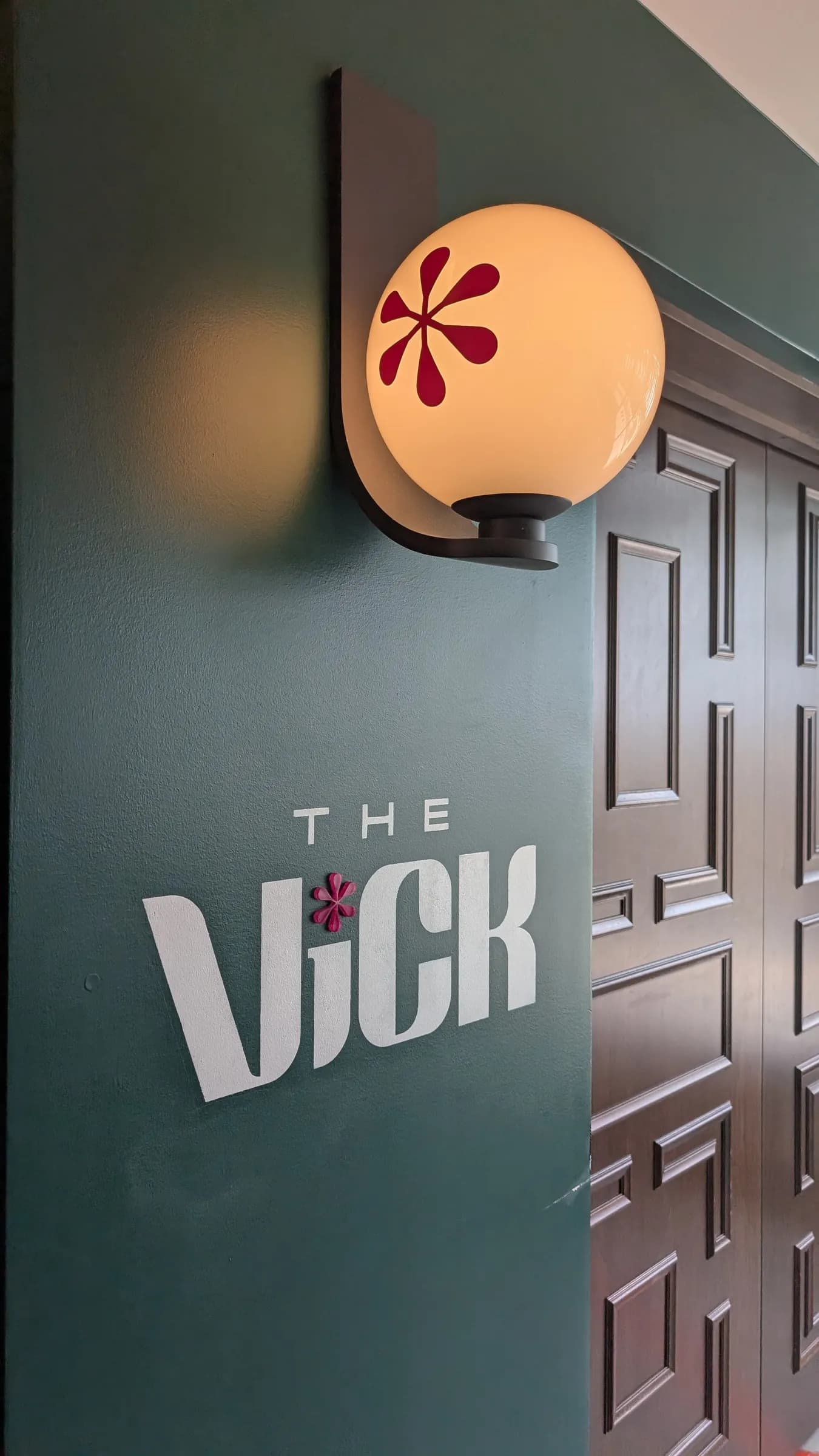 The sign for The Vick, the rooftop bar at Hotel Celare: white letters on a deep green wall, under a glowing globe lamp painted with the same red asterisk that dots the i