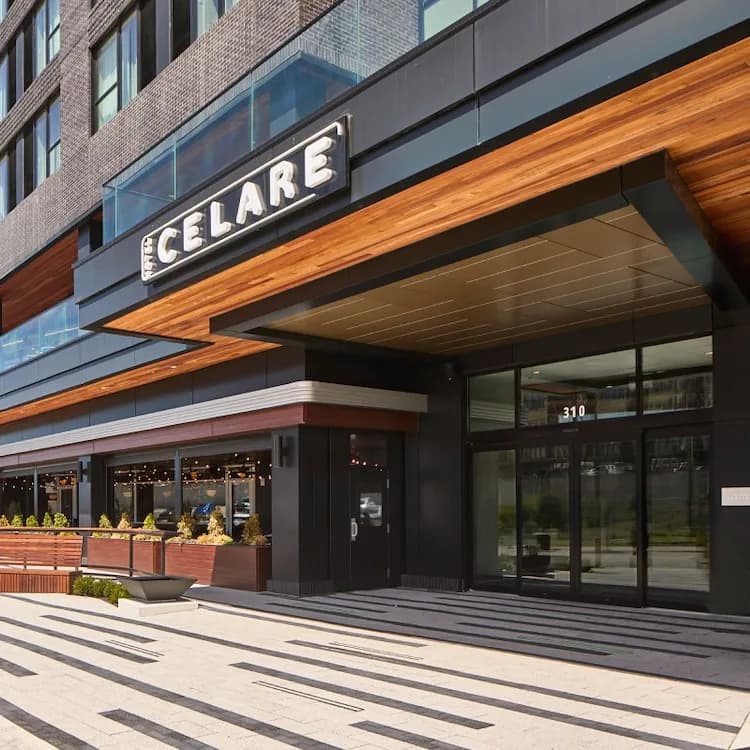 The entrance to Hotel Celare, with its name on the canopy over the glass doors and a striped plaza with wooden benches out front